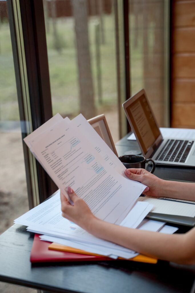 pexels photo 9222427 9222427 Person holding and reviewing documents near window with laptops on a desk.